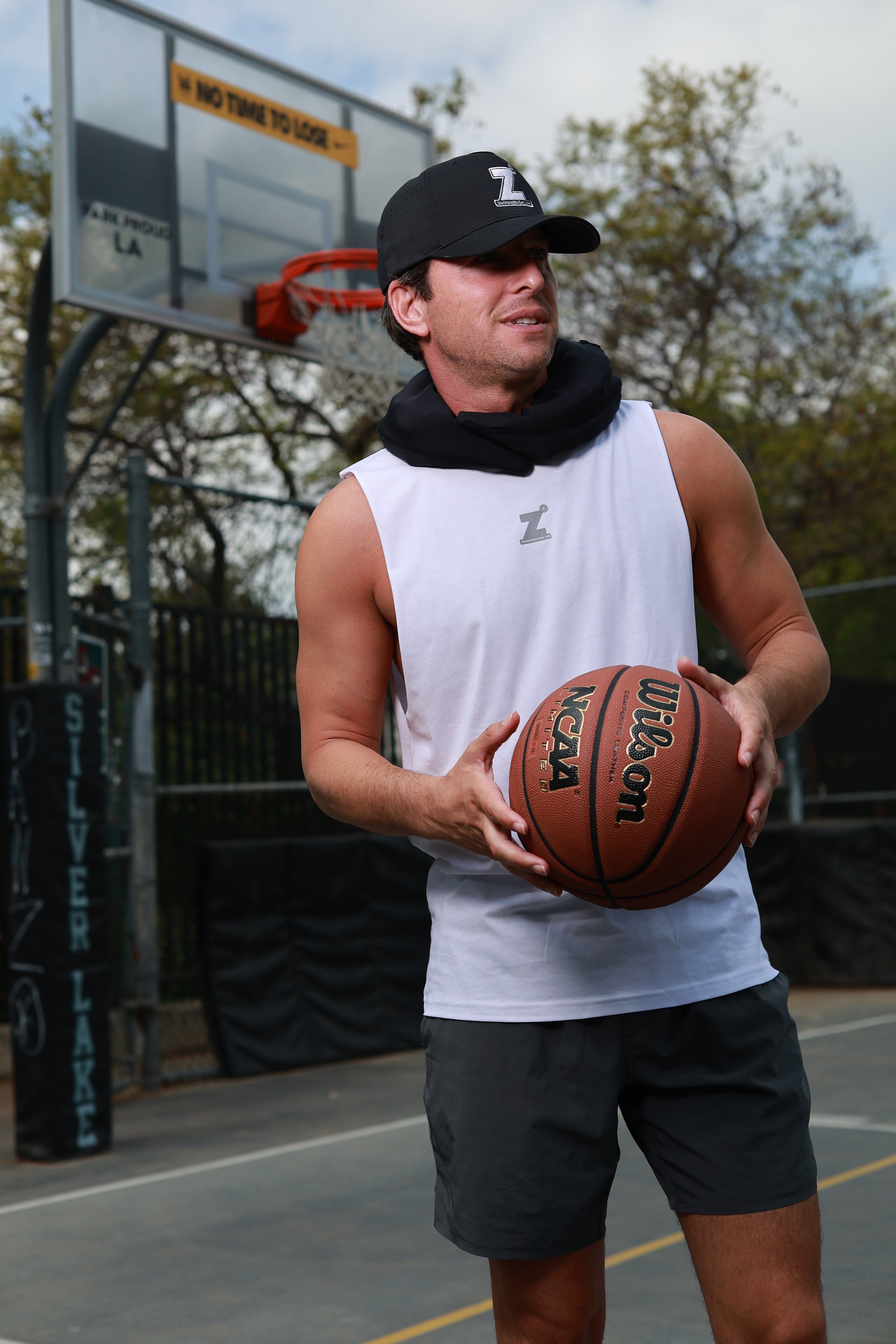 Man holding a basketball on an outdoor court with a black cap and white sleeveless shirt. He has a HERO around his neck