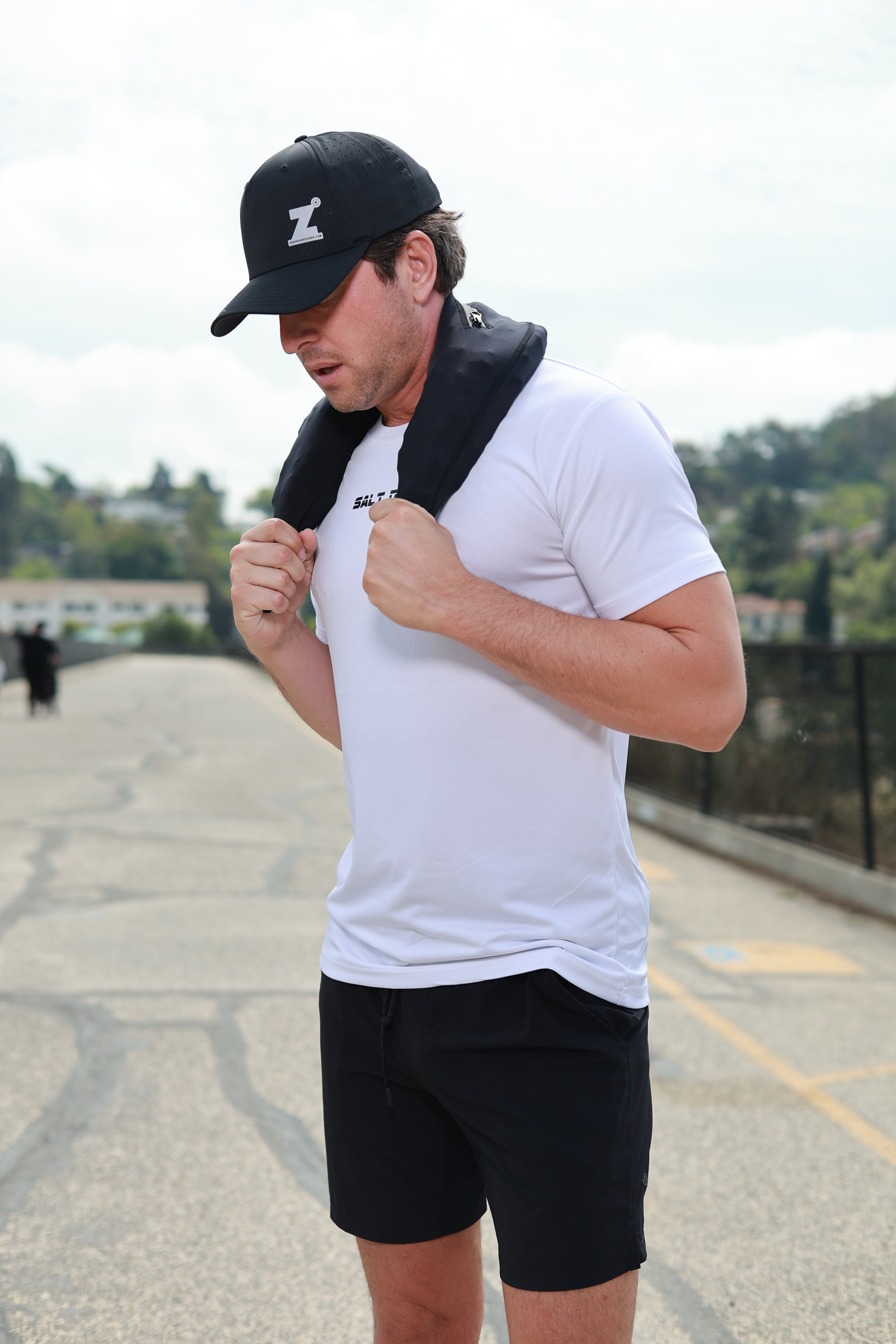 Man wearing a white t-shirt, black shorts, and a black cap with black neoprene pouch on his neck, standing on a street.