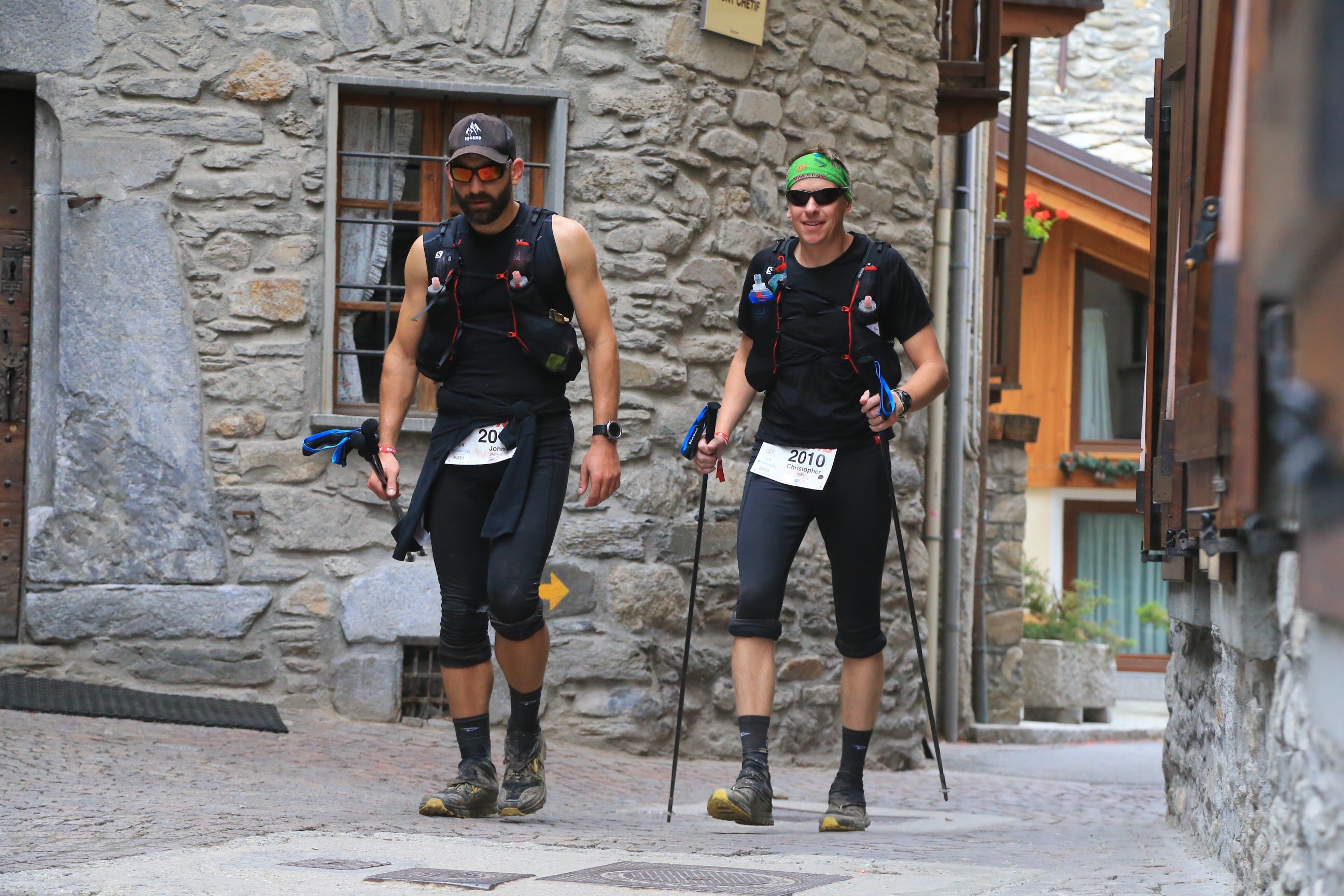 Two athletes with backpacks and running gear walking through a village street.